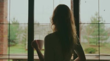 Elinde bir fincan sıcak doğal çay, elinde kahve olan mutlu genç bir kadın içkinin tadını çıkarıyor. Güzel kız rahat bir odada dikilip pencereye bakıyor. Yeşil arka plan. Normal kot pantolon..
