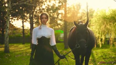 Atı olan seksi bir kadın. Model kız güçlü yüzlü at elle tutulan evcil hayvanla yürüyor. Peri Kraliçesi siyah uzun elbise eski tarz. Günbatımı orman ağaçları sihirli sıcak güneş ışığı 4k.