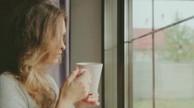 Sarışın sakallı genç bir kadın elinde bir fincan sıcak çay, elinde taze öğütülmüş kahve, içmekten zevk alıyor. Yetişkin kızlar için rahat bir oda. Pencere kenarına bakıyor. Gri giysi 4K.