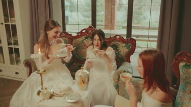 Sıcak çay içen mutlu bir grup kadın ellerinde fincanlarla altın sette. Asil Bayan Neşeli Surat. Retro kanepe sehpası. Kız dinleniyor çay partisi veriyor kraliyet konforlu oda beyaz eski moda elbise.