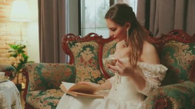 Sıcak çay içen mutlu kadın elinde fincanlar kağıt roman okuyor. Sarışın kadın asil bayan güzel yüz dudakları. Seksi kız kanepede dinlenir kraliyet odası beyaz klasik elbise eski tarz