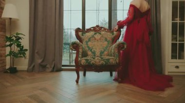 Sanat kraliçesi eski tarz taht işçilerinin kumaşa dokunduğu yerde dolaşıyor. Uzun kırmızı elbise treninin arkası. Kraliyet sandalyesi beyaz oda penceresi. Seksi prenses masalı Gotik kadın nedime..