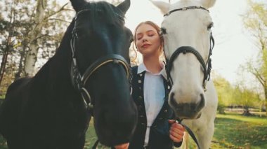 Çift atlı güzel yüzlü fantezi kadın. Genç Jokey kız ahırda iki atın dizginlerini tutuyor. Siyah beyaz bir hayvan. Aşk evcil hayvanı. Yaz doğası. Güneş ışığı. 4k ışık parlaması.