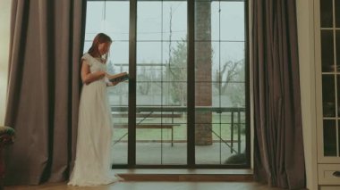 Pencerede el ele tutuşup kitap okuyan genç bir kadın. Yetişkin güzel kız dudakları. Kraliyet konforlu odasında dinlenin. Beyaz kaliteli elbise. Uzun balo elbisesi. 4k.