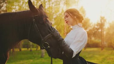 Atlı mutlu kadın fantezisi. Gülümseyen güzel kız, güzel surat, dudaklar, sevgi dolu elleriyle atına sarılıyor, ağızlık takıyor. Peri masalı kraliçesi siyah uzun elbiseli eski tarz orman güneşi ışığı 4k