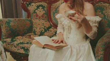 Sıcak çay içen mutlu kadın elinde fincanlar kağıt roman okuyor. Asil bayan güzel yüz dudaklarını yakından çek. Geriye dönük seksi kız kanepede dinlenir kraliyet odası beyaz klasik elbise eski usul. 4k.