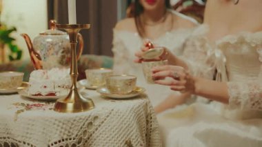 Kadın şeker kasesini elinde tutuyor ve çay kaşığıyla sıcak çay fincanı setine şeker koyuyor. Kanepede oturan asil bayan kahve masası kızı oturma odasında dinleniyor. Eski tarz beyaz elbise..