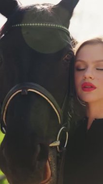 Kapalı portre güzellik yüzü kırmızı dudaklar mavi gözler mutlu kadın at eli ile ağızlık siyah kürke dokunuyor, dikkat et at evcil hayvanı. Sarışın, seksi kız. Sihirli güneş ışığı. dikey video kırpma kısa.