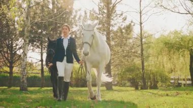 Güzel yüzlü genç bir kadın at mankeni ile yürüyor. Mutlu Jokey kız ahırda iki atı dizginliyor siyah ve beyaz hayvan. Aşk evcil hayvanı. Yaz doğası güneş ışığı parlıyor. 4k