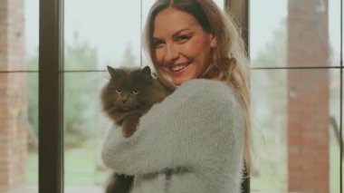 Mutlu sarışın kadın pencerenin kenarında durmuş elinde güzel tüylü gri kediyi tutuyor, neşeli kız sevgiyle bakıyor, gülen surat hayvan ağızlık kürkünü öpüyor. Konforlu ev kıyafetleri