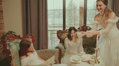 Mutlu, gülümseyen bir kadın çaydanlığa sıcak çay doldurur altın, zarif bardak setine, kahve masasındaki kanepede oturan asil kadınlar çay içer. Oturma odasındaki dinlenen bayan beyaz, eski tarz elbise.