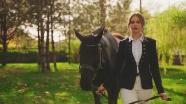 Güzel yüzlü, geçmişe dönük, mutlu, atıyla yürüyen kadın. Parlak güneş ışığı yeşil çimen orman parkı ağacını aydınlatıyor. Jokey giysili kız prenses eski usul para, dizginler siyah at. Aşkım.