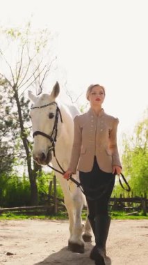 Güzel, sarışın, mutlu, at yürüyüşü yapan, yaz güneşi yeşil çimen ağacı olan bir kadın. Jokey kıyafetleri içinde yetişkin bir kız elinde siyah dizginler beyaz at, evcil hayvanı sevmek için dikey video