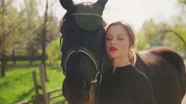 Yakın plan güzellik, kırmızı dudaklar mavi gözler at eli ile mutlu kadın ağızlık siyah kürke dokunuyor dostluk bakımı at hayvanı. Havalı seksi kız kucaklaşması eşofmanı. Sihirli güneş ışığı ağacı 4k