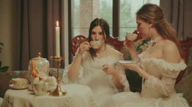 Sıcak çay içen mutlu bir grup kadın ellerinde fincanlarla altın sette. Asil Bayan Neşeli Surat. Retro kanepe sehpası. Kız dinleniyor çay partisi veriyor kraliyet konforlu oda beyaz eski moda elbise.