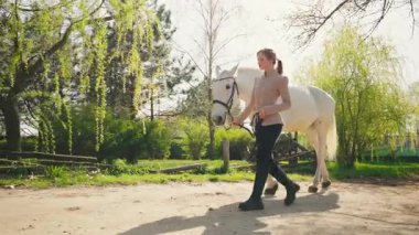 Güzel, sarışın, mutlu, at yürüyüşü yapan genç bir kadın. Yaz güneşi yeşil çimen ağacı toprak yolunu aydınlatıyor. Jokey elbiseli yetişkin kız elinde siyah dizginler beyaz at, konsept aşk hayvanı