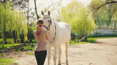 Güzel, sarışın, mutlu, at kuyruğu manzaralı kadın. Yaz güneşi yeşil çimen ağacını aydınlatıyor. Jokey gibi giyinmiş yetişkin bir kız. El, beyaz at başı ağızlık, konsept, sevgi, hayvan bakımı.