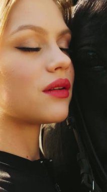 Kapalı portre güzellik yüzü kırmızı dudaklar mavi gözler mutlu kadın at eli ile ağızlık siyah kürke dokunuyor, dikkat et at evcil hayvanı. Şık, seksi kız. Sihirli güneş ışığı. ağaç dikey video kırpma kısa.