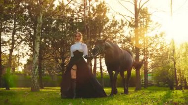 Atı olan seksi bir kadın. Model kız güçlü yüzlü at elle tutulan evcil hayvanla yürüyor. Peri Kraliçesi siyah uzun elbise eski tarz. Günbatımı orman ağaçları sihirli sıcak güneş ışığı 4k.