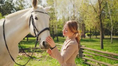 Güzel, sarışın, atıyla mutlu bir kadın. Yaz doğası güneşi yeşil çimen günü. Gülümseyen yüz, okşayan el öpen beyaz at kafası ağızlık, konsept aşk bakımı hayvan evcil hayvanı. Jokey elbiseli kız.