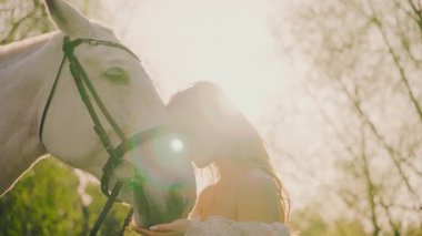 Atı olan seksi mutlu kadın. Güzel yüzlü manken sarışın kız beyaz atına sevgiyle sarılıyor. Peri Kraliçesi 'nin ağızlık evcil hayvanı. Orman günbatımı ağaçları sihirli güneş gökyüzünü aydınlatır 4k