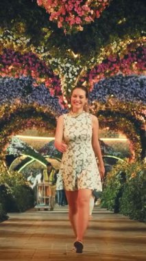 Mutlu kadın turist, Dubai 'nin çiçek açan bahçesinde romantik bir akşam aralığı boyunca yürüyor. Dans eden girdaplar. Gülen surat. Peyzaj tasarımı çiçekler kalp şeklinde kemerler. Gece çelengi aydınlatması