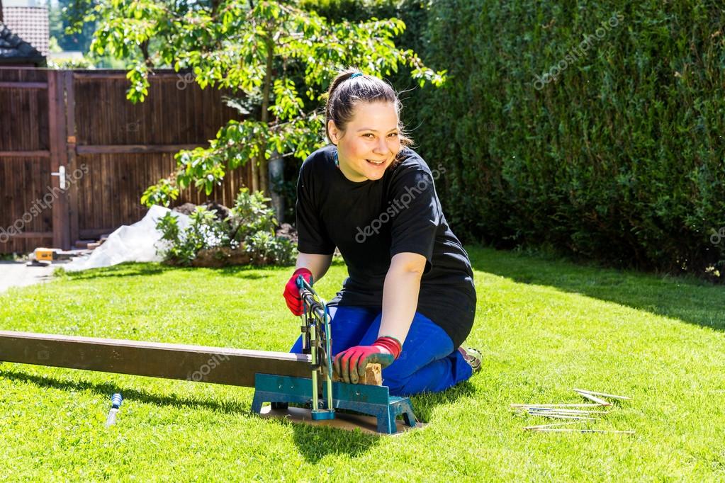 Mädchen sägt in Arbeitskleidung einen Holzbalken im Garten