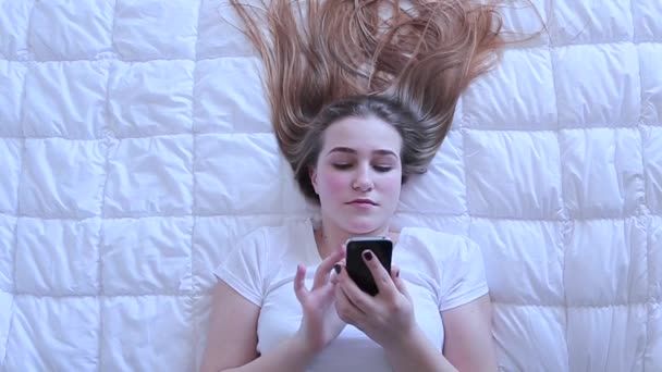 Дівчина друкує повідомлення на мобільному пристрої, що лежить на ліжку зверху — стокове відео