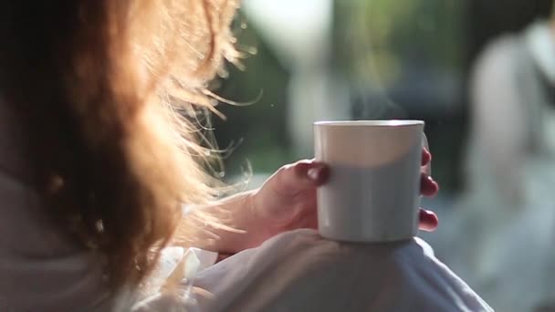 Chica se sienta en un abrigo blanco en la cama con un café — Vídeos de Stock