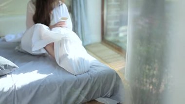 Bir fincan kahve ile beyaz bir yatakta kız oturuyor