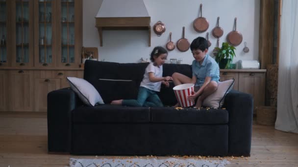 Kleines Mädchen mit einem Jungen bestreut Popcorn auf dem Sofa — Stockvideo