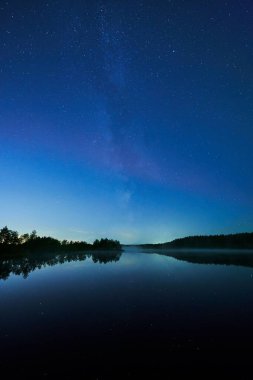 Sakin göl yüzeyine yansıyan yıldızlarla dolu koyu mavi bir gece göğü.