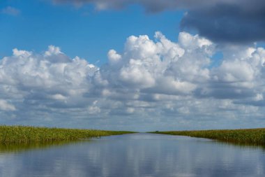 Sakin suyun manzarası mavi gökyüzünü ve yeşil çimenler arasındaki bulutları yansıtıyor.
