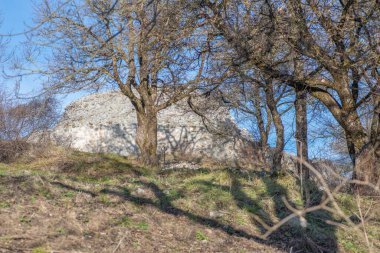 Çıplak ağaçların arkasında beliren büyük beyaz kaya oluşumu güneşli yamaçlara gölge düşürüyor..