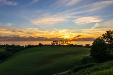 Renkli bulutlar ve yeşil yamaçlarla okyanusun üzerinde canlı bir günbatımı.