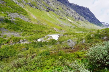 Küçük bir deresi olan yemyeşil dağ vadisi ve parlak bir gökyüzünün altındaki sarp kayalık yamaçlar boyunca eriyen kar parçaları..
