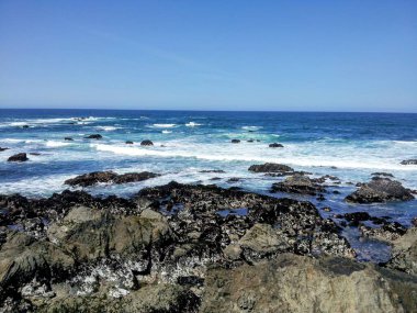 Dalgalar mavi gökyüzünün altında kayalık kıyılara çarpıyor.