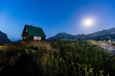 Ay ışığında dağlarla çevrili bir kulübe.