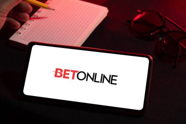 Bogota, Kolombiya - 25 Kasım 2025: Bet Online logosu ekranda gözlüklü bir masada dinlenen cep telefonu; arka planda bulanık bir el deftere yazıyor. Spor bahisleri ve dijital platformlar kavramı