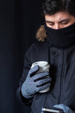 Kışlık ceket, eldiven ve eşarp giyen bir adam elinde sıcak kahve, soğuk havada günlük rutin bir konsept tutarken telefonunu kontrol ediyor.