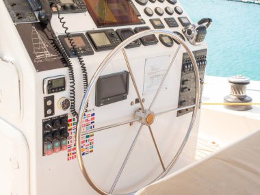 Bir yatın kokpit kontrollerinin parlak, temiz bir görüntüsü, VHF radyo, çeşitli deniz bayrağı çıkartmaları ve sakin mavi okyanusa bakan büyük metal bir dümen..