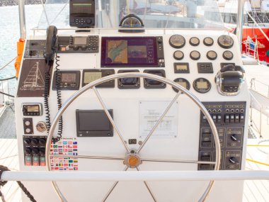 Modern bir tekne miğferinin detaylı bir görüntüsü, büyük paslanmaz çelik bir tekerlek, bir sürü gösterge, bir VHF radyo, navigasyon ekranları, bir dizi bayrak çıkartması.