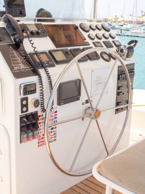 Bir yatın kokpit kontrollerinin parlak, temiz bir görüntüsü, VHF radyo, çeşitli deniz bayrağı çıkartmaları ve sakin mavi okyanusa bakan büyük metal bir dümen..