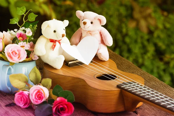 Ukulele 'de iki ayı, oyuncak ayı ve güller Eski ahşabın üzerinde beyaz kağıt kalp kartıyla