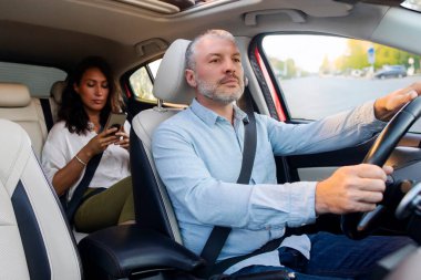 Confident man taxi driver driving car with passenger woman on backseat using cellphone. Cab transportation concept