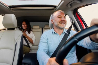 Taxi service concept. Happy man driver with wireless earphones driving car, holding hands on steering wheel and smiling, woman passenger sitting on backseat
