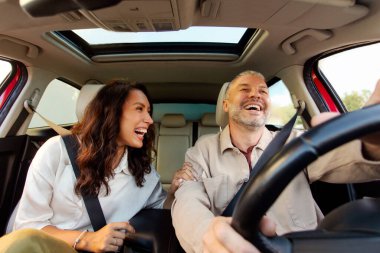 Enjoying fun travel. Overjoyed European couple sitting on the front passenger seats, talking and laughing, man driving car, view from dashboard