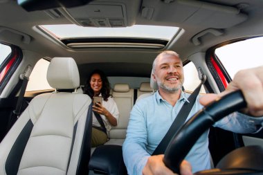 Cheerful middle aged male driver riding car and smiling, lady passenger sitting on backseat. Modern transportation and taxi service concept