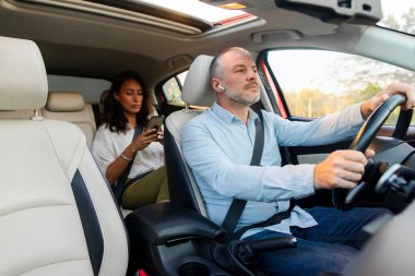 Man driver with wireless earphones driving car, woman passenger sitting on back seat and using cellphone. Transport, taxi service and modern technologies concept