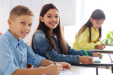 Two friends classmates, school girl and boy sitting at desks and smiling at camera during lesson, enjoying learning atmosphere in private school